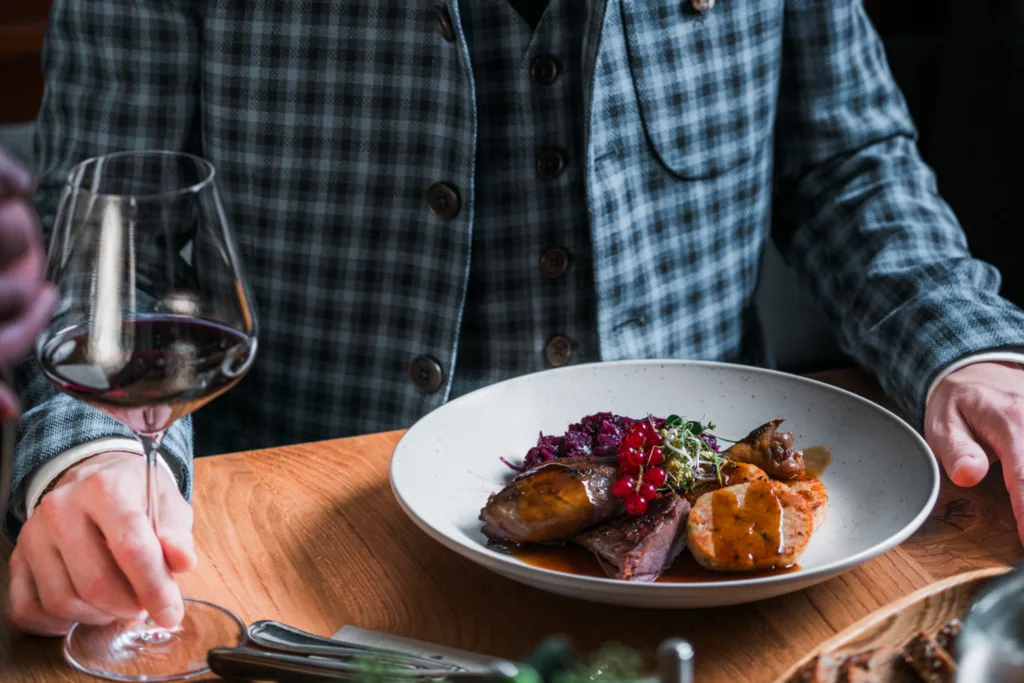 Ein Mann im blau-schwarzkarrierten Sakko sitzt vor einem gebratenen Gansl mit Rotkraut und Semmelknödeln. In der Hand hält er ein Glas Rotwein.