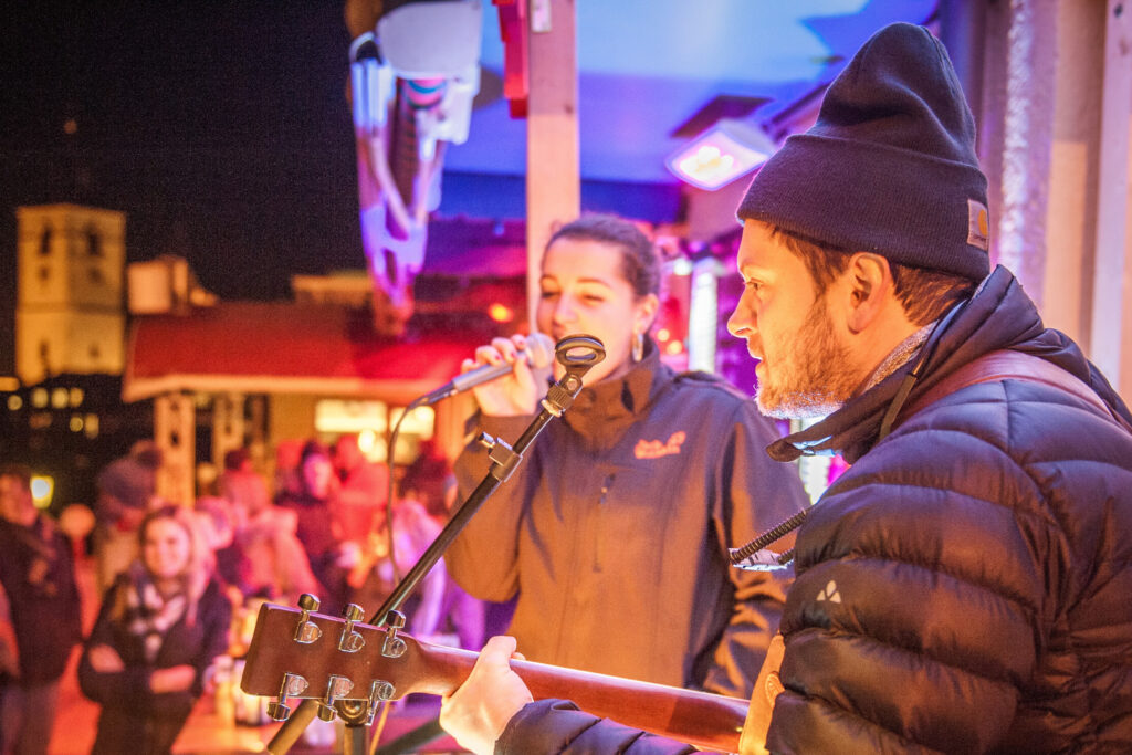 Ein Gitarrenspieler in dicker Jacke und eine Sängerin, die in ihr Mikrofon singt. Sie befinden sich auf der Winter-Seeterrasse von Paul der Wirt. Im Hintergrund der Kirchturm.