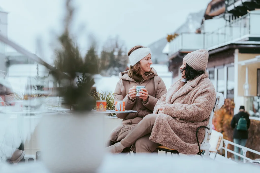 Zwei Freundinnen auf der winterlichen Seeterrasse von Paul der Wirt. Eine hält eine Tasse mit Punsch und blickt nach hinten.