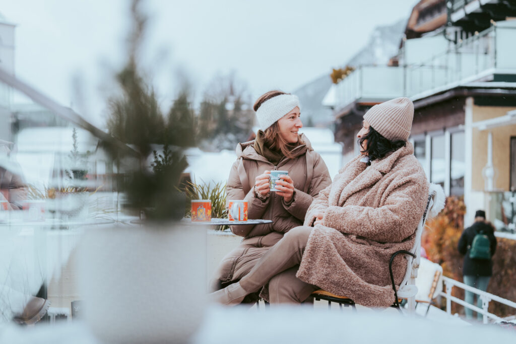 Zwei Freundinnen auf der winterlichen Seeterrasse von Paul der Wirt. Eine hält eine Tasse mit Punsch und blickt nach hinten.